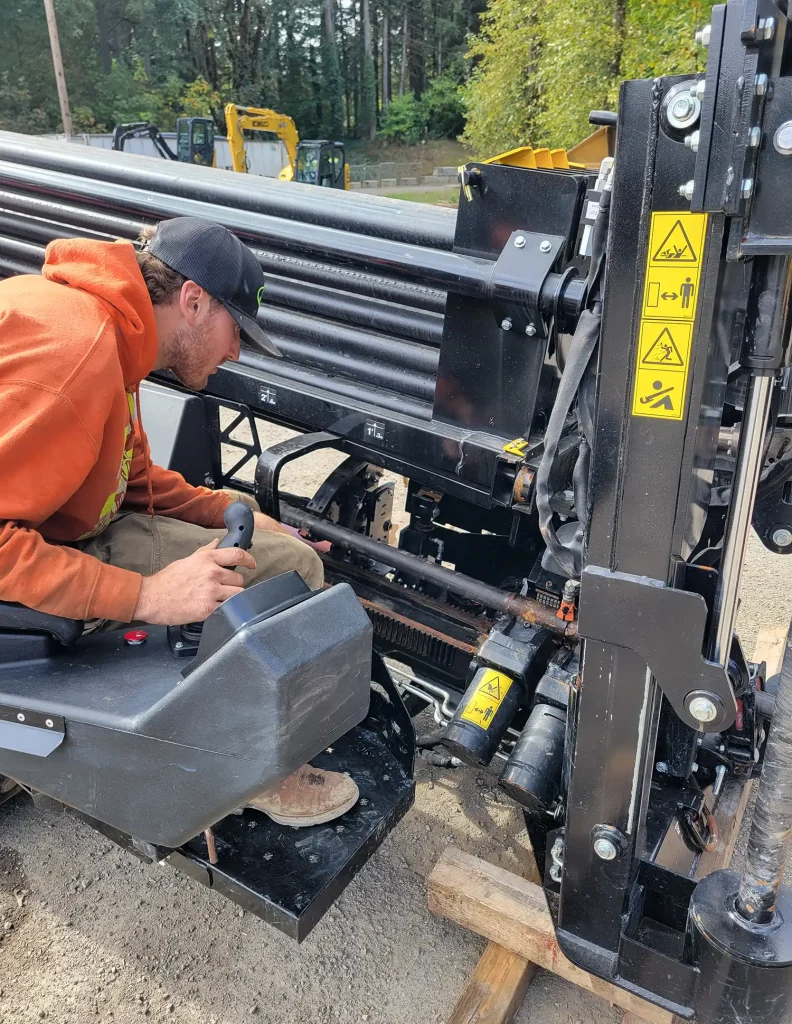A worker repairs machinery, focusing on the hydraulic components while seated on a heavy-duty equipment base.