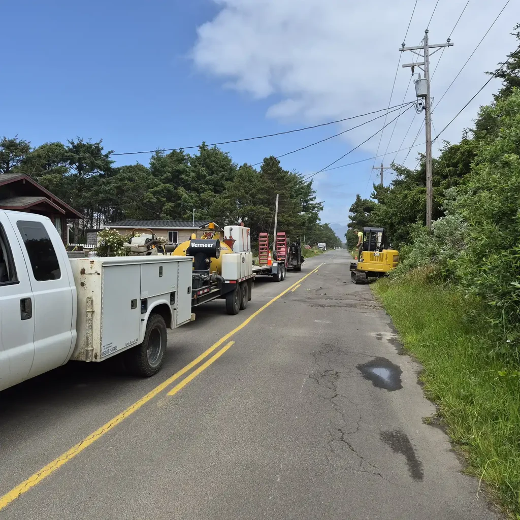 Construction vehicles are parked along a narrow road surrounded by trees under a clear blue sky.