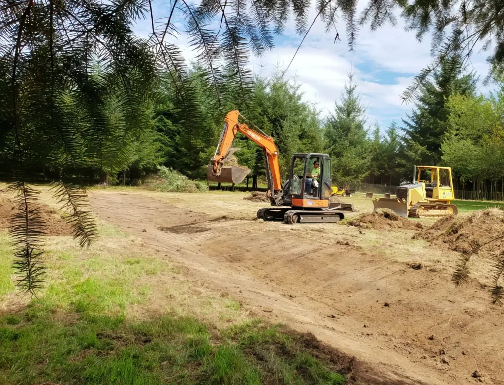 Excavators and heavy machinery work on a cleared construction site surrounded by trees and open land.