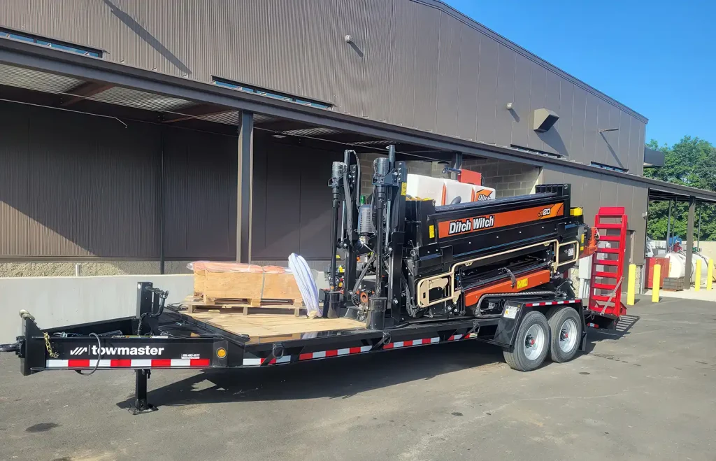 A loaded trailer with construction equipment, including a large machine and wooden pallets, parked outside a warehouse.