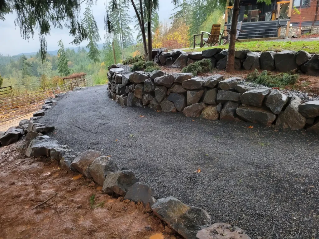 Curved gravel pathway bordered by stone walls, leading through a wooded area with a view of the landscape.