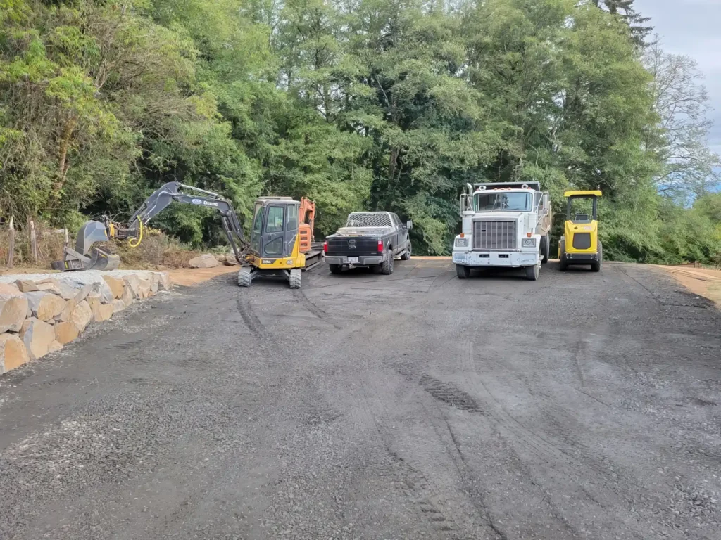 Construction vehicles, including an excavator and a dump truck, are parked on a gravel site surrounded by trees.