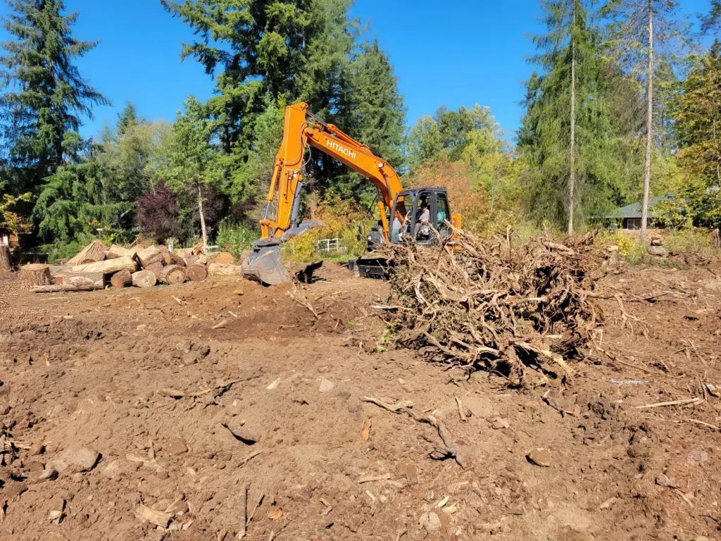 Excavator clearing land with a large pile of branches and dirt under a clear blue sky surrounded by trees.